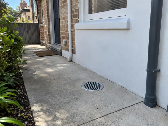 Concrete side passage with a round drain grate beside brick and rendered walls in Crows Nest